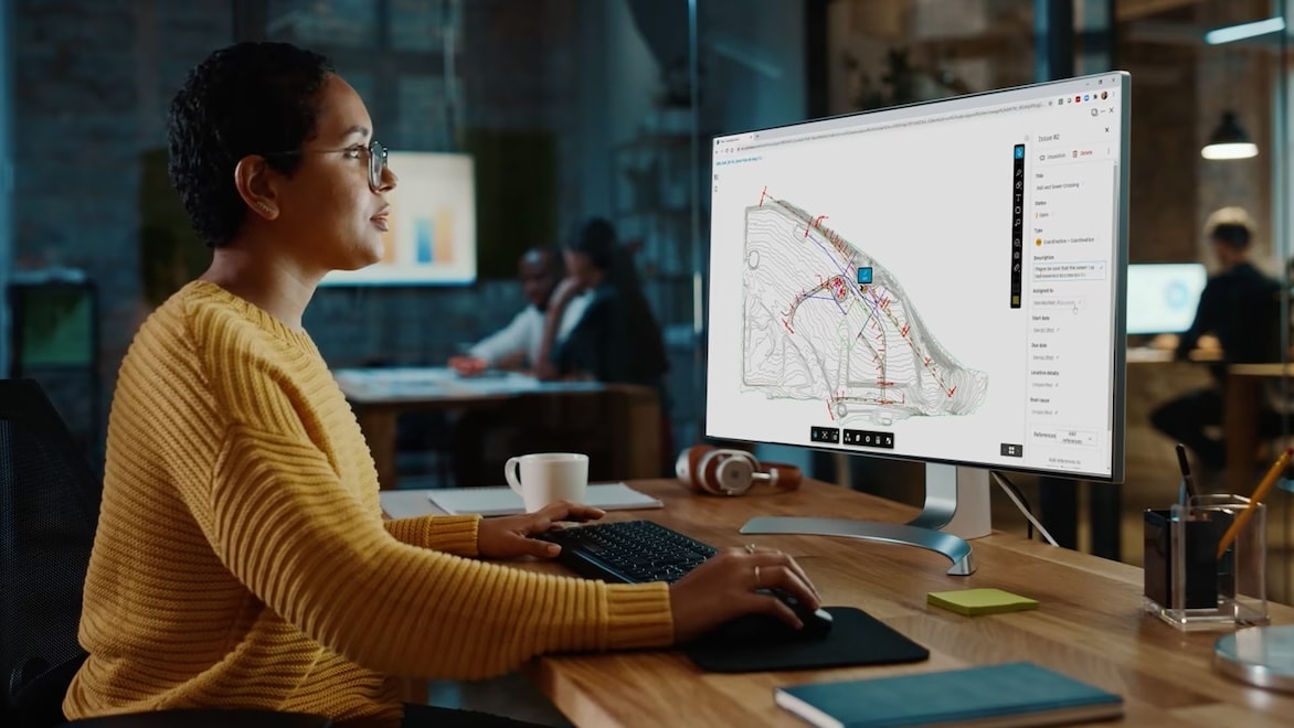 A woman in an orange sweater sits at a computer displaying a building model rendering.