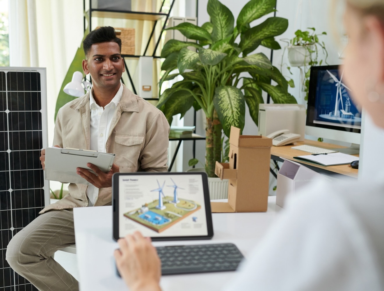 Two workers in an office consult a sustainable building project on a laptop. A solar panel is in the background.