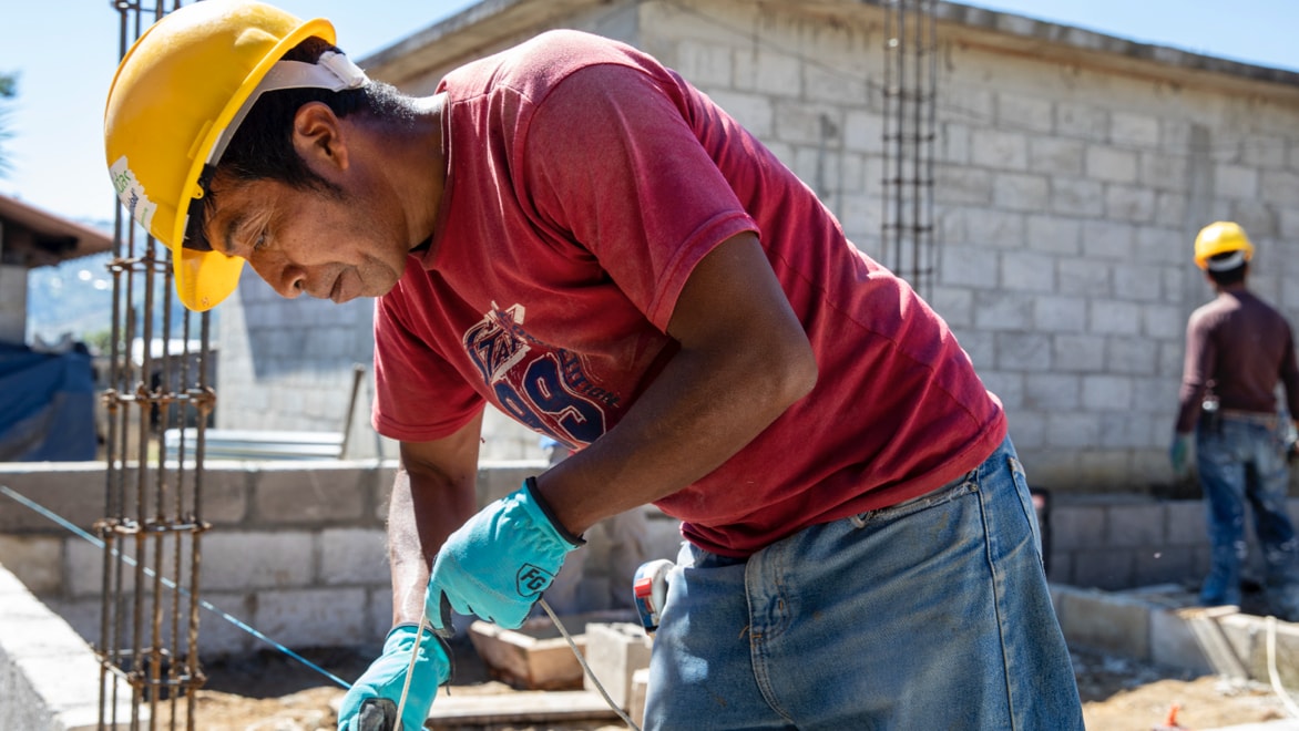 A construction worker wearing a red graphic t-shirt, yellow hard hat, work gloves, and blue jeans, leaning over a wall made of concrete blocks, pulling on a string or wire connected, looking down at the task at hand. Background: sunny sky, a further-developed concrete block wall, and another worker wearing the same attire looking away from the camera at the constructed wall.