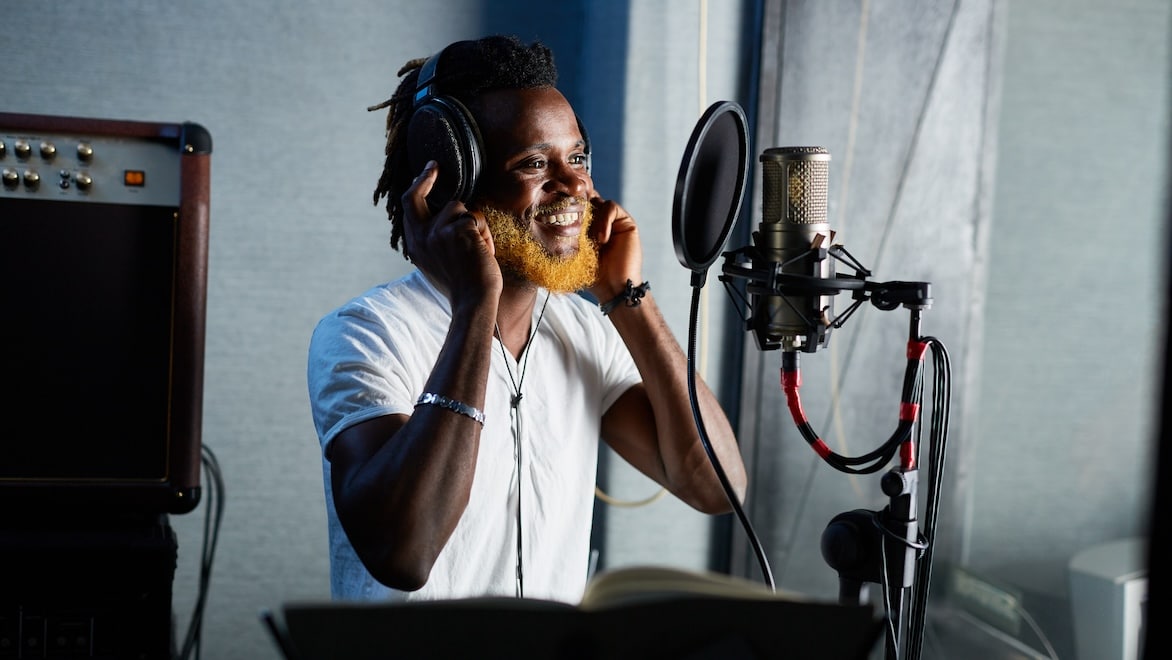 A young man wearing headphones stands in front of a microphone in a recording studio.