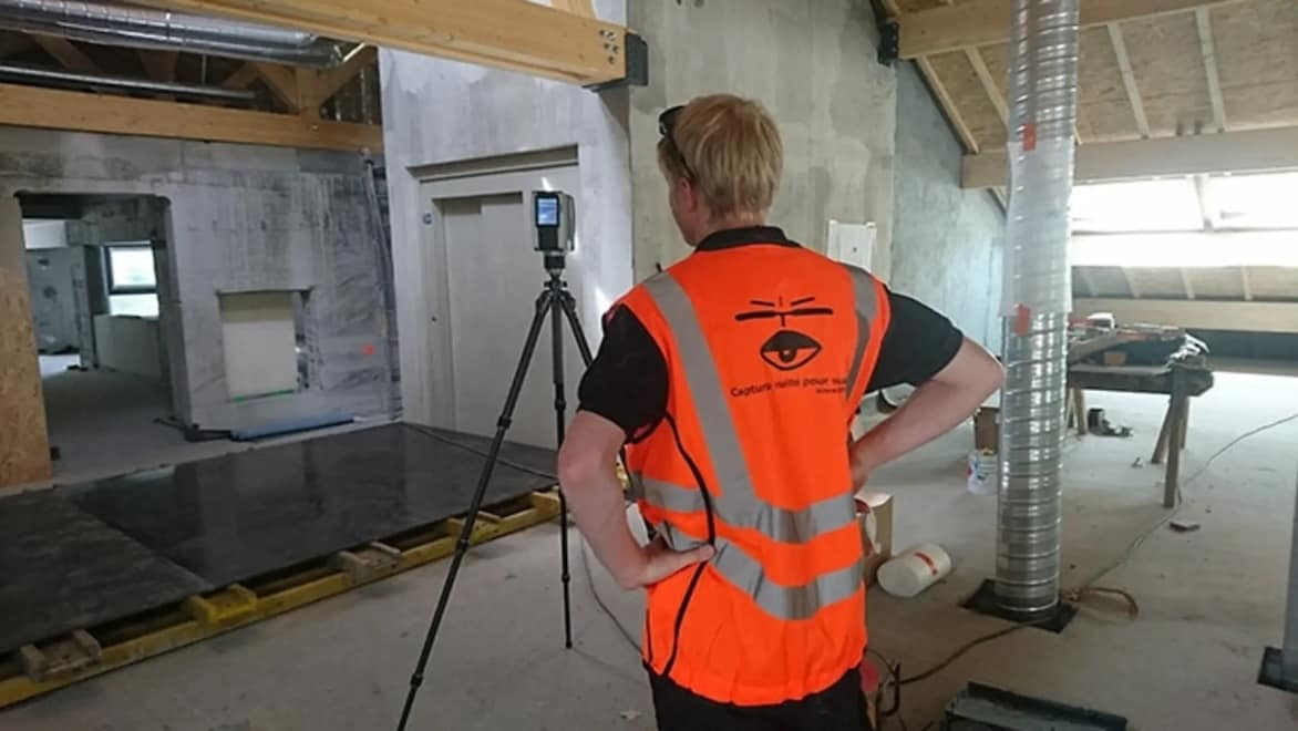 A surveyor in an orange vest stands next to a point cloud scanner on a tripod while scanning the interior of an incomplete construction project.