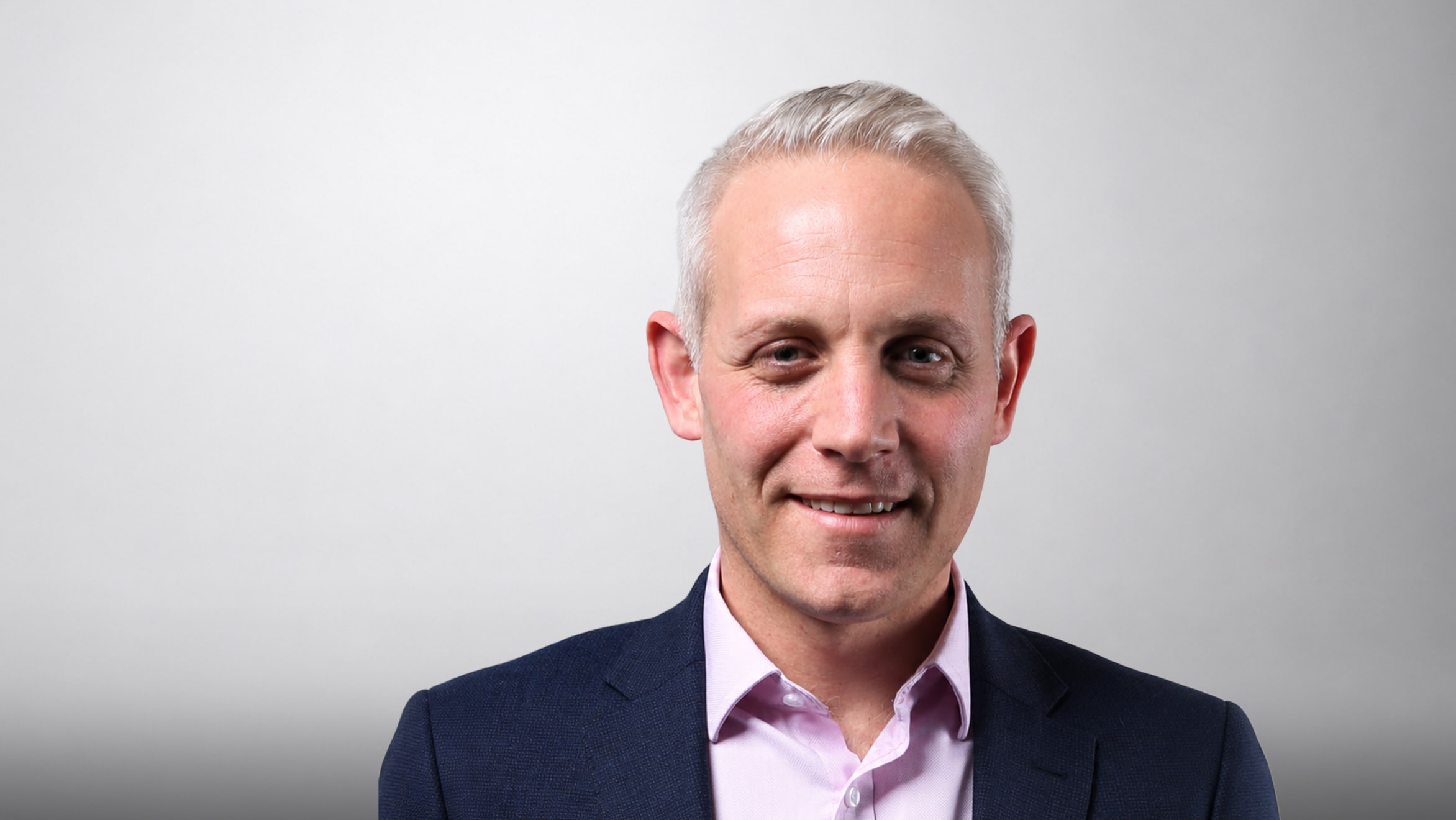 Portrait of a smiling man with grey hair in a suit against a light background.