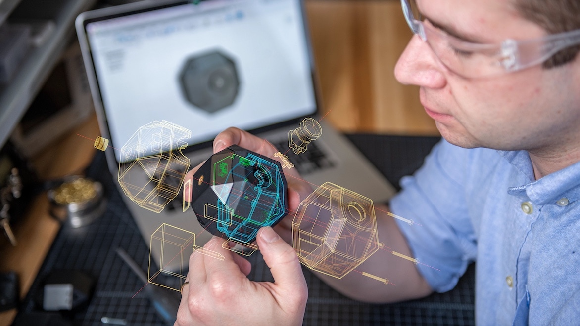 A man holds a machine part prototype next to a computer screen displaying its design elements.