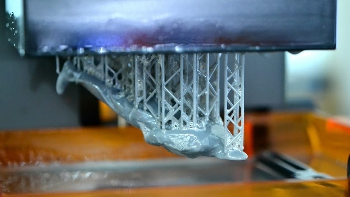 A close-up of a figurine being produced on an SLA printer shows the still-wet liquid polymer and scaffolding.