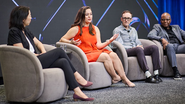 Four people sitting in armchairs on a stage with a dark background, one woman in an orange dress speaking with hands gesturing