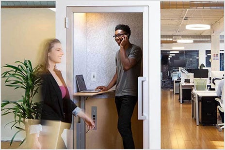 A man stands inside a compact office booth speaking on the phone while a blurred woman carrying a laptop walks past.