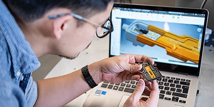 A man in the Autodesk Technology Center examining an orange and black computer chip over his laptop.  