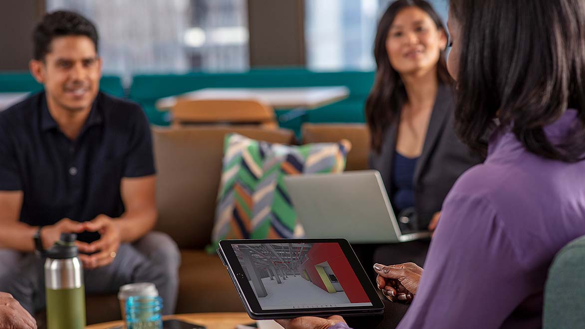 A group of people is seated in a casual meeting setting, discussing a project displayed on a screen. The screen shows a 3D architectural model, while other devices are also visible, indicating a collaborative work environment.
