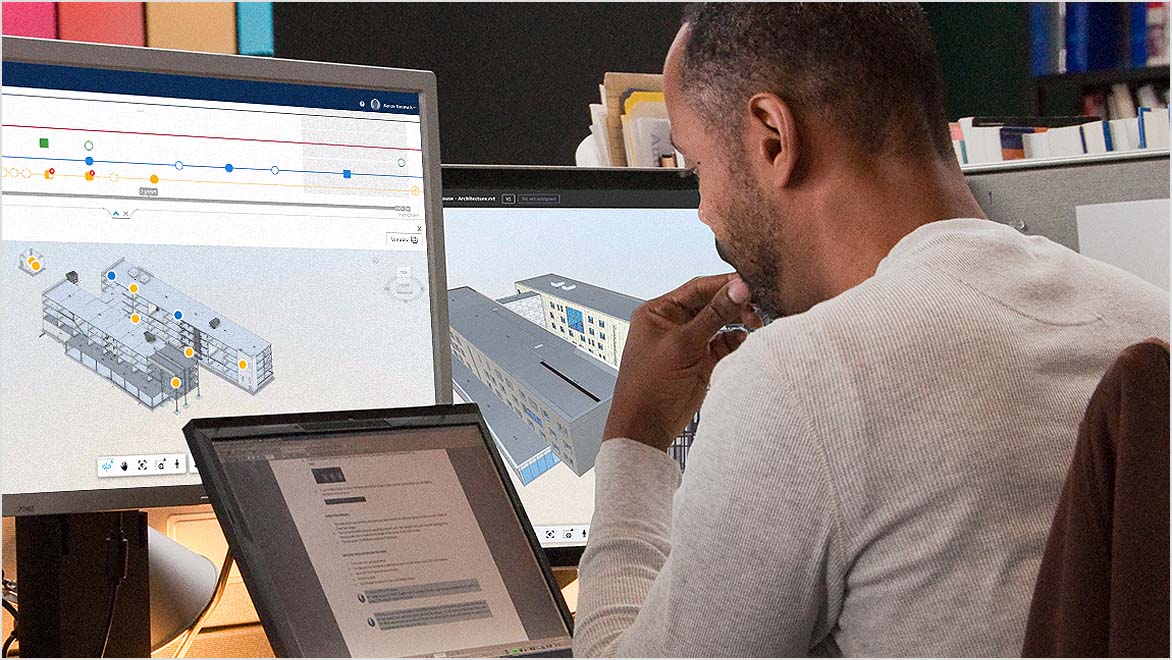 A person is seated at a desk, viewing two computer screens that display architectural models and project data within the Autodesk for Government Cloud environment.