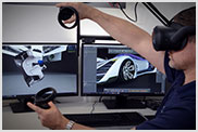 Person wearing a VR headset and holding controllers, in front of two monitors displaying a car design