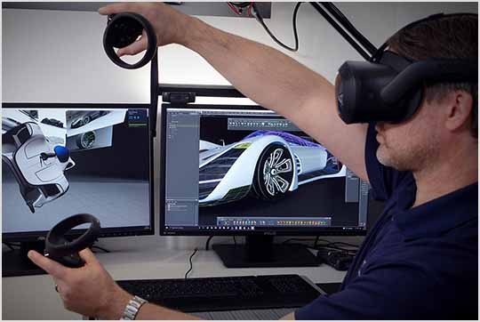 Person wearing a VR headset and holding controllers, in front of two monitors displaying a car design