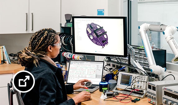 Eine Ingenieurin hält ein Bauteil in der Hand und betrachtet am Bildschirm das gleiche Bauteil in der EAGLE-Oberfläche für den Elektronikentwurf.