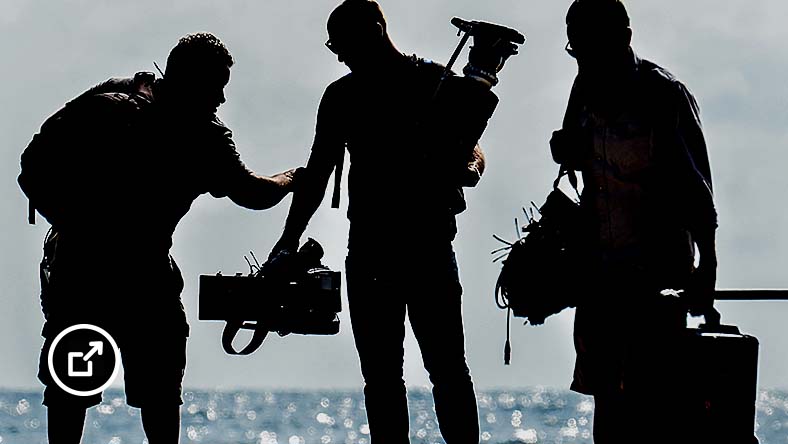 Silhouettes of three members of a film crew standing on beach