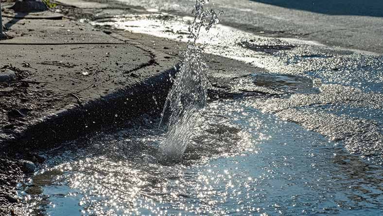 A burst of water erupts from a break in a concrete sidewalk, creating a splash