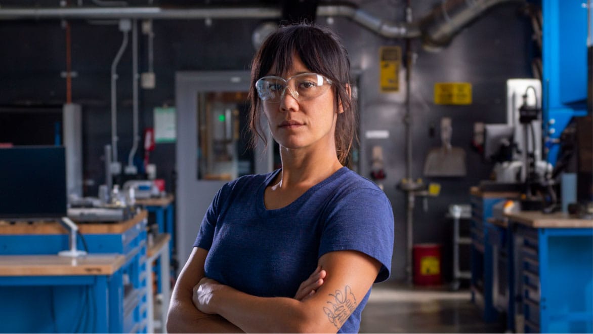 woman wearing safety glasses and standing in a machining shop while looking at the camera with arms crossed.