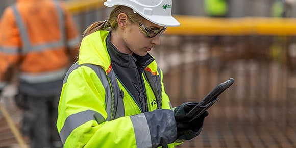 A woman in a white hardhat and yellow jacket using a tablet on a jobsite 
