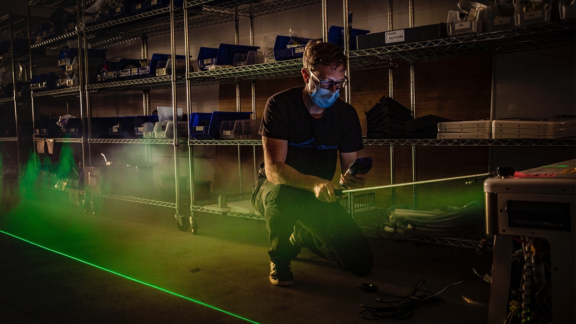 Person wearing mask kneels in a manufacturing shop with green light beam and steam