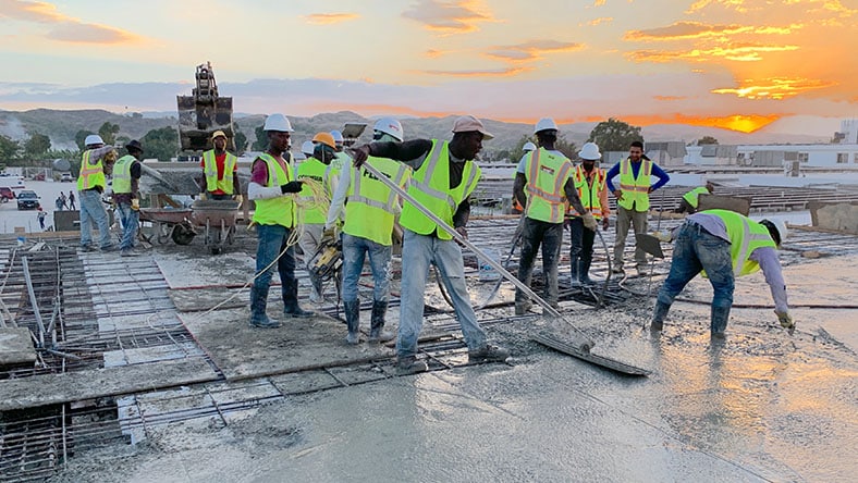 Group of workers wearing hi-viz yellow jackets pouring and smoothing cement for a Build Health International construction project, against a backdrop of other single-story white buildings and a glowing orange/yellow sunset.