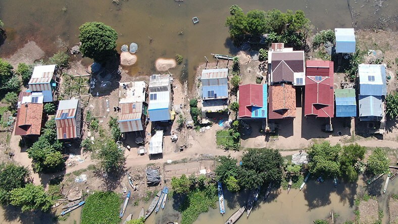 Aerial view of a group of small houses with rooftop solar panels, along the shoreline of a lake surrounded by trees.