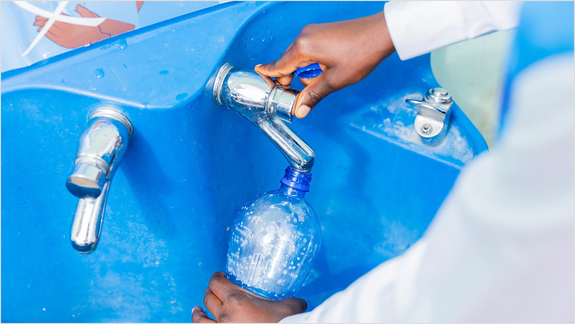 Hands of a child pushing a faucet button to fill a clear plastic water bottle at a blue water station.
