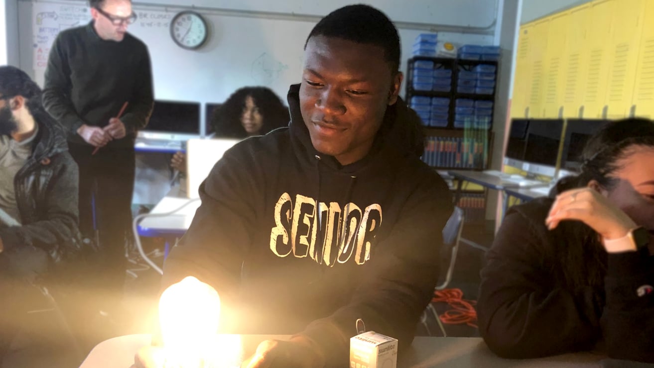 Student sitting in classroom wearing "Senior" hoodie, looking at lightbulb project as part of Stacks+Joules training. Instructor standing in background talking to another student.