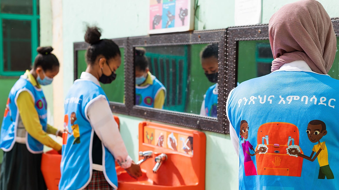 Three young women students wearing light-blue Splash-branded vests washing their hands at three Splash handwashing stations along a wall in a school.