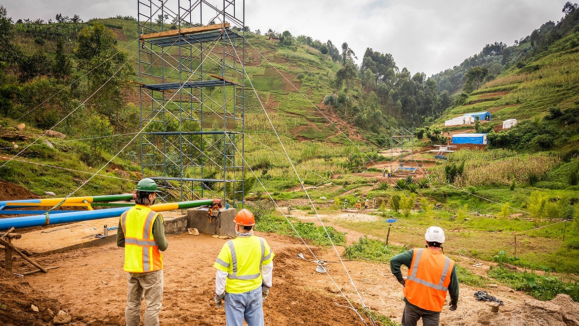 Three construction workers wearing hi-viz yellow and orange vests and hard hats look out on a B2P bridge under construction, with a four-story scaffolding supported by guy-lines. Green grassy hills and colorful bridge materials in the distance.