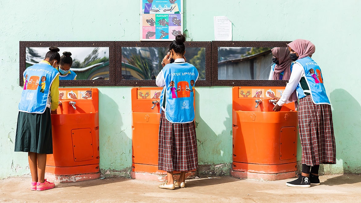 Three young women students wearing light-blue Splash-branded vests and long skirts, washing their hands at three Splash handwashing stations along a wall in a school.