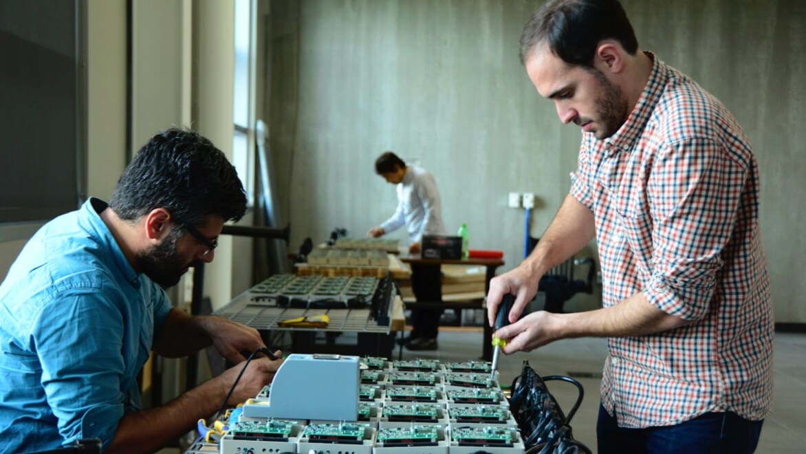 Foreground: Two men working on electrical boards. Background: Another person working on a separate set of electric boxes.