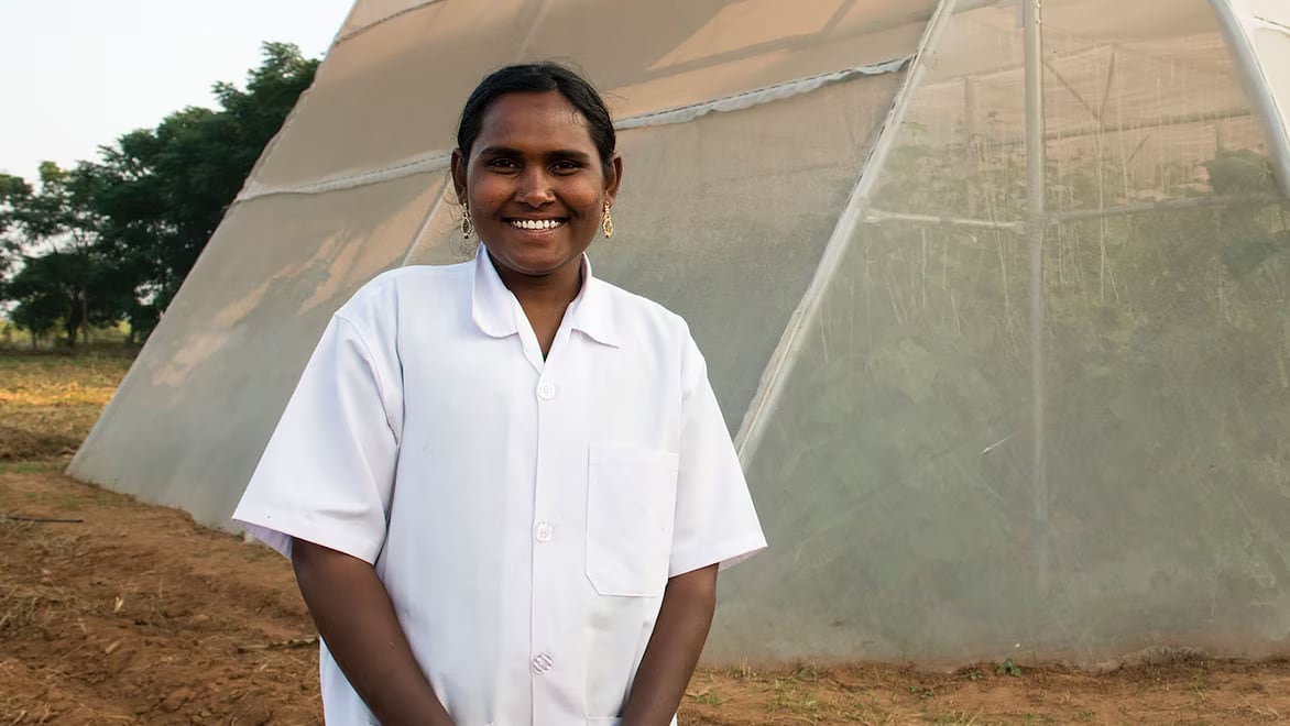 An Indian woman smallholder farm stands in front of one of Kheyti's "Greenhouse-in-a-Box" greenhouses on a sunny day, smiling and looking at the camera, wearing a white button-down t-shirt and gold-colored earrings. Grass surrounds the greenhouse; tall green trees in the background.