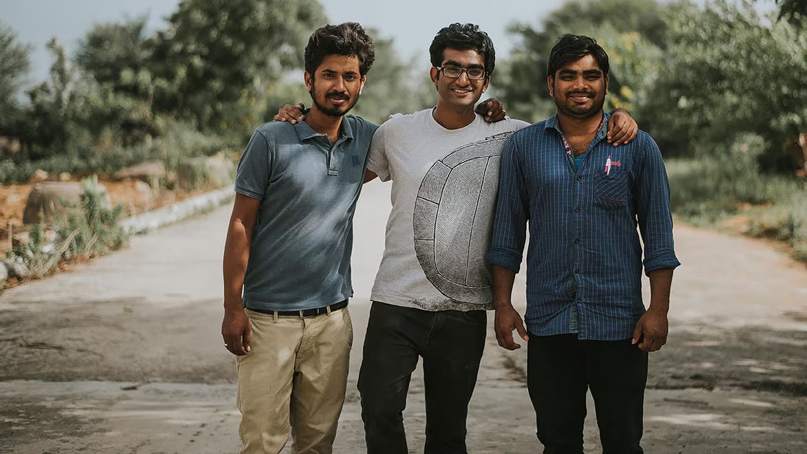 Three young Indian men standing on a street on a sunny but somewhat overcast day, smiling and posing for a group photo. The two men on the right and in the middle are wearing dark jeans; the one on the left is wearing khakis and a blue-grey collared t-shirt. The man in the middle is wearing a white t-shirt; the man on the right is wearing a dark blue, striped button-down shirt with rolled-up sleeves. Trees and vegetation line the road in the background.