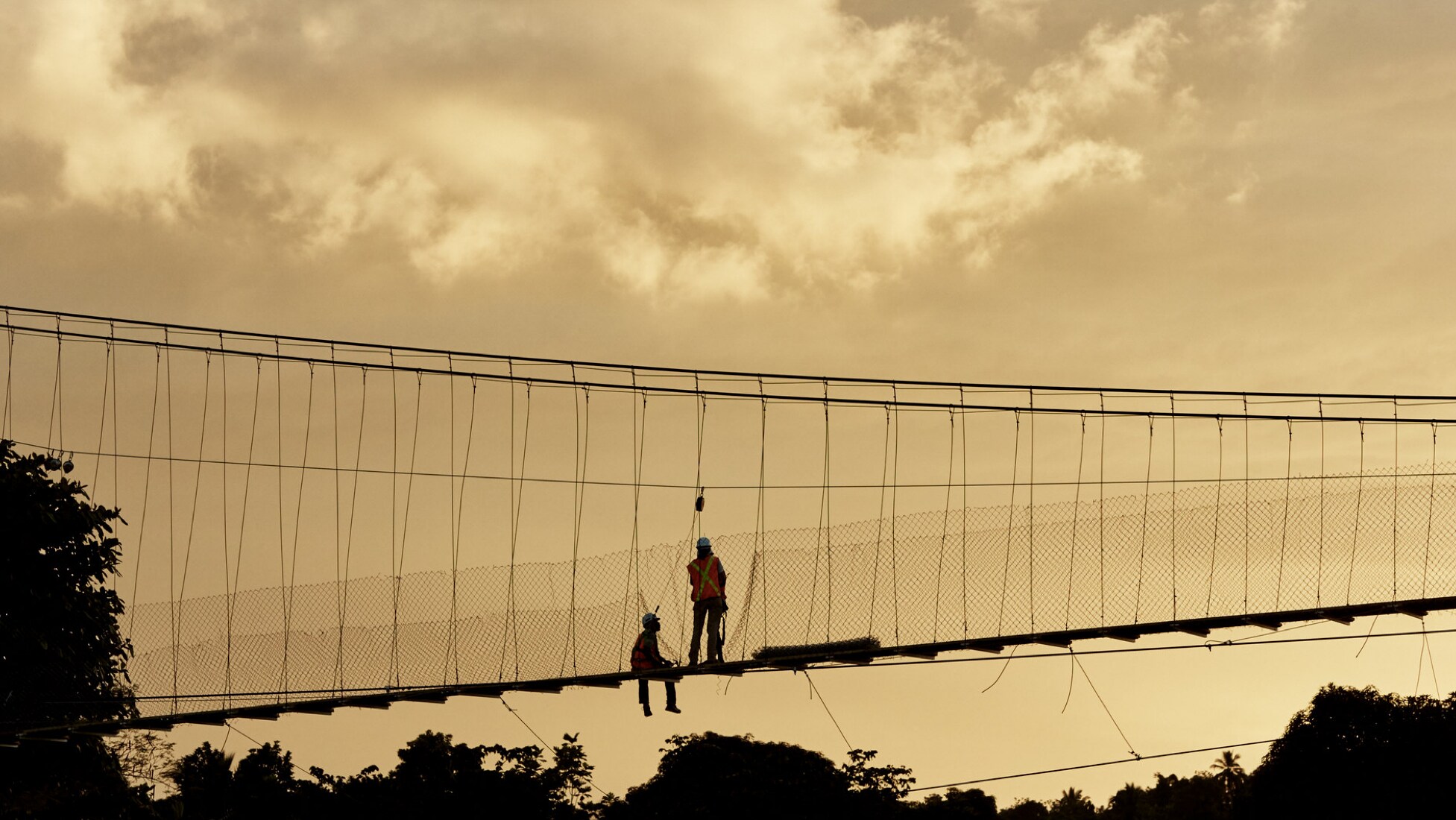 A distant side-view photo of a B2P suspension bridge with two construction workers on the bridge—one sitting with his legs hanging off the edge, the other standing up next to the one sitting—silhouetted by the glow of an open sky at dusk.. 