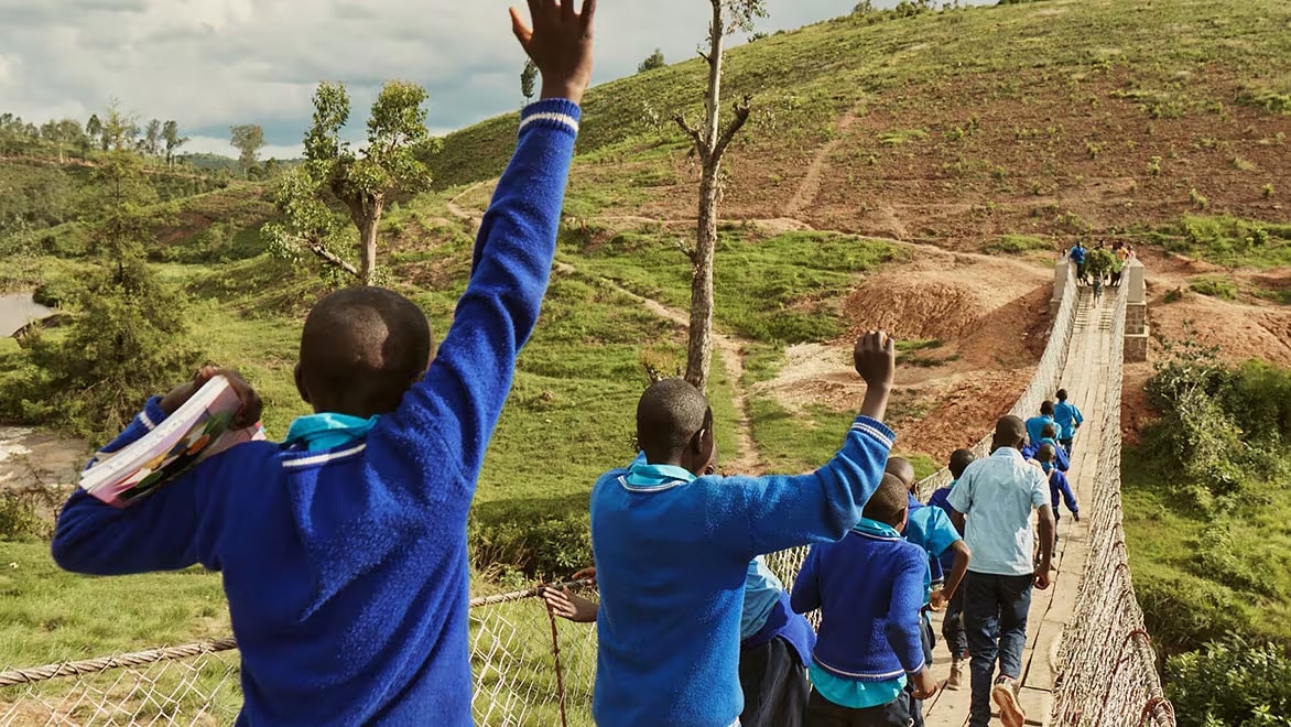 Wide angle shot of ~10 school children wearing blue sweaters walking over a B2P trailbridge in Rwanda. A teacher wearing a light-colored, button-down t-shirt accompanies the children on the bridge.