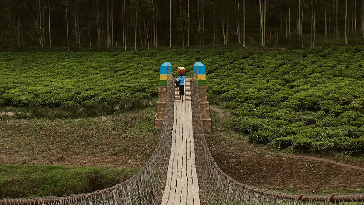 First-person view of a B2P trailbridge from one end looking to the other. The other end of the A person carrying something on their head, standing at the bridge is surrounded by lush green shrubs. Green grass and hilly terrain on either side of the trailbridge. An individual carrying something under their left arm, holding something else on top of their head with their right hand, walking across the bridge, almost to the other/far side.