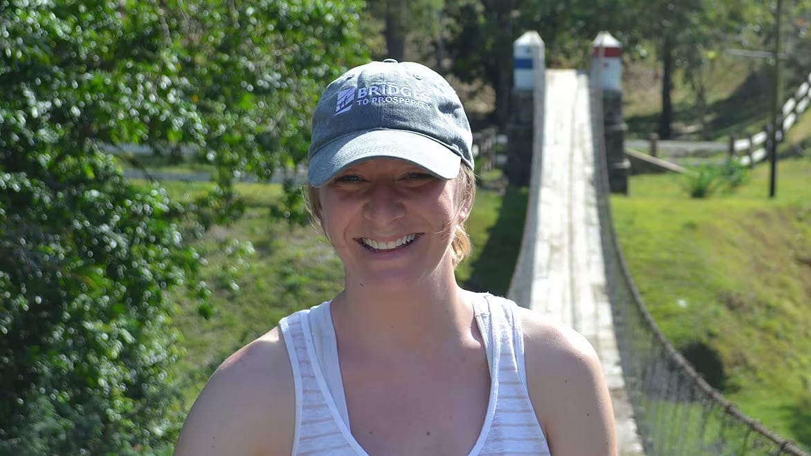 Headshot of Alissa Davis, B2P Director of Business Development, standing at one end of a B2P trailbridge, wearing a grey B2P hat, smiling and looking at the camera. The bridge is surrounded by hilly terrain covered with green grass.