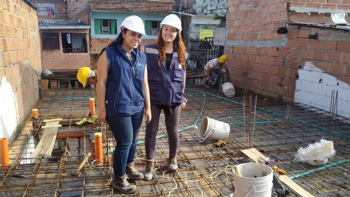 Two women Build Change workers wearing white hard hats smiling at the camera for a group shot, standing on the rebar framing of a roof at a Build Change renovation project.