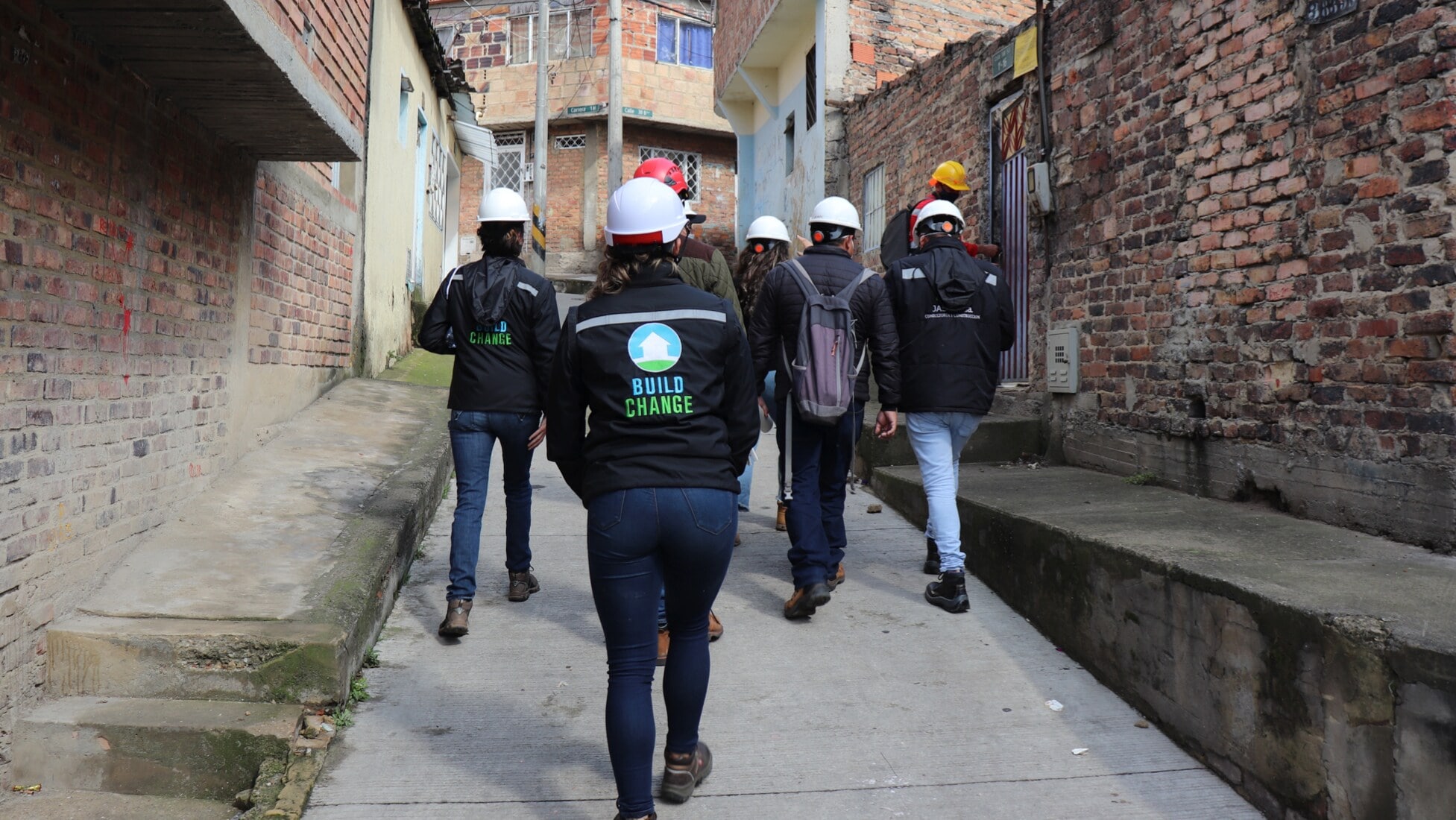 Six individuals with their backs turned to the camera walking down a narrow alleyway in Colombia, with brick walls on either side of them. All individuals are wearing hard hats and jeans; three are wearing Build Change-branded jackets.