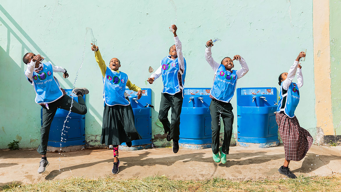 Five young students wearing light-blue Splash-branded vests, jumping and smiling, playing with water in front of four blue Splash handwashing stations along an exterior wall at a school.